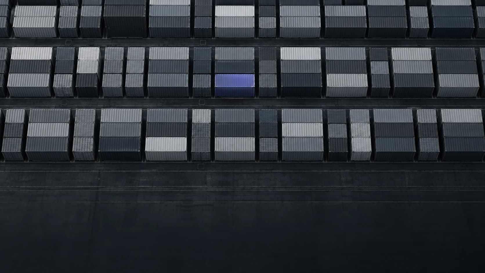 Aerial view of stacked shipping containers in rows at a terminal, with one container lit in violet